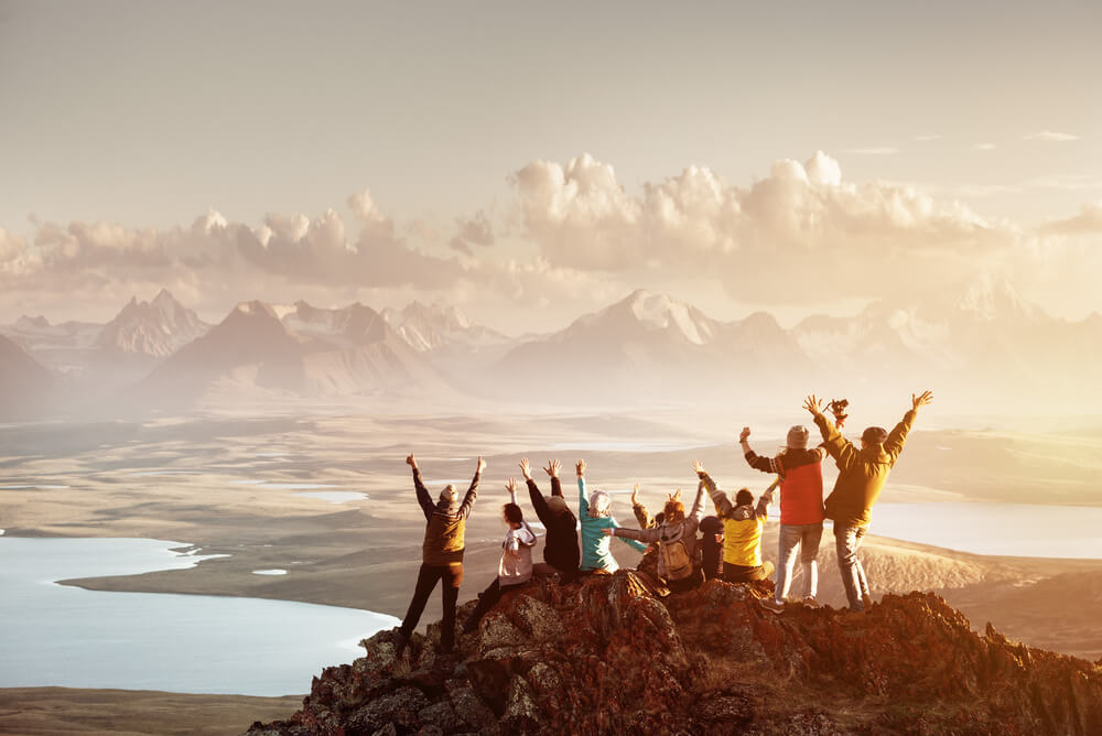 A big group of friends celebrating on the top of a mountain against a backdrop of sunsets, lakes and mountains